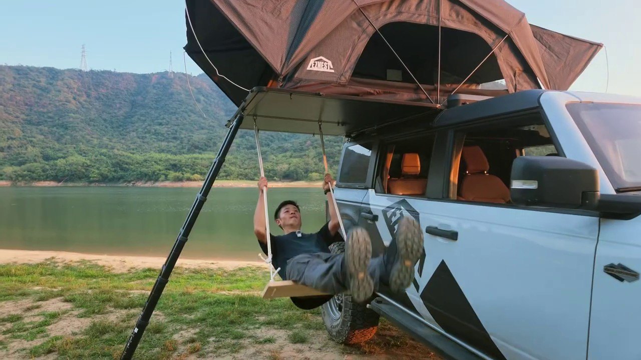 Open aluminium roof tent on a white SUV beside a lake, with a swing hung beneath it